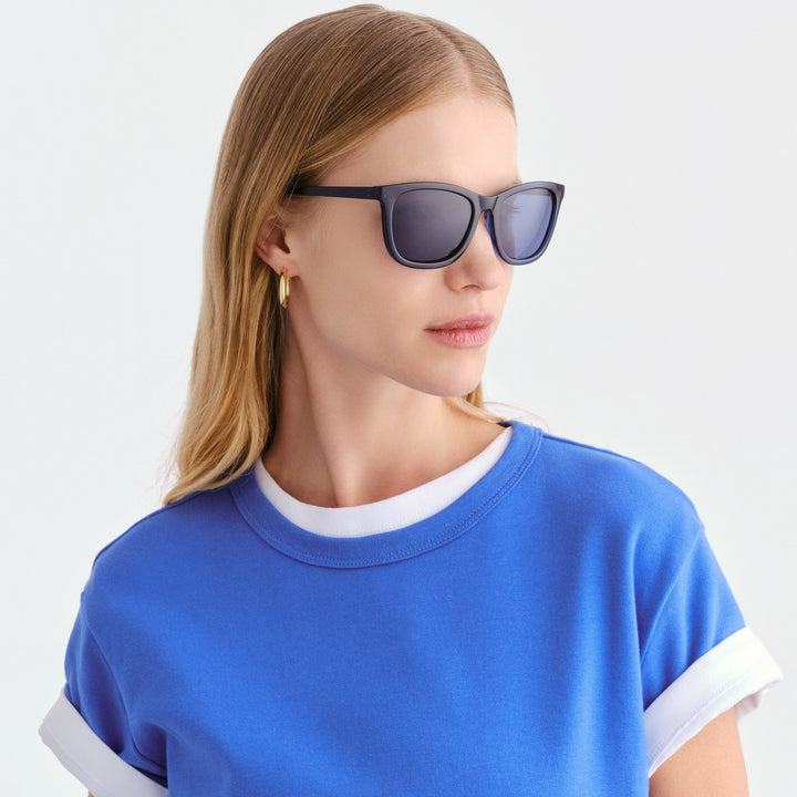A woman with blonde hair wearing navy sunglasses with dark lenses and a blue ringer t-shirt, photographed against a clean white background.