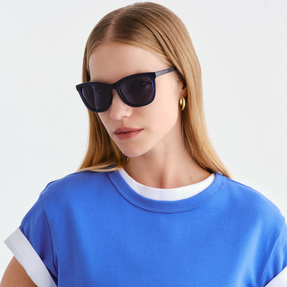 A woman with blonde hair wearing black cat-eye sunglasses and a blue ringer t-shirt with white trim, photographed against a clean white background.