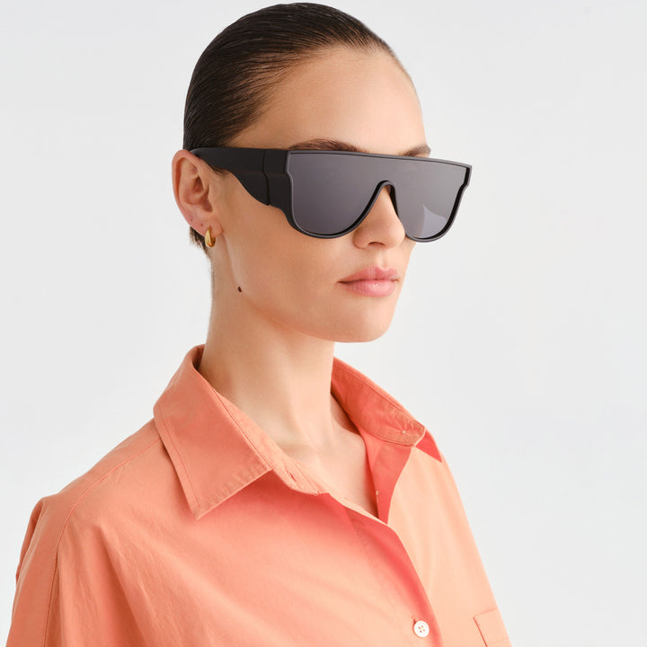 A woman in profile wearing futuristic black shield-style sunglasses with dark lenses and a peach-colored collared shirt, against a light gray studio background.
