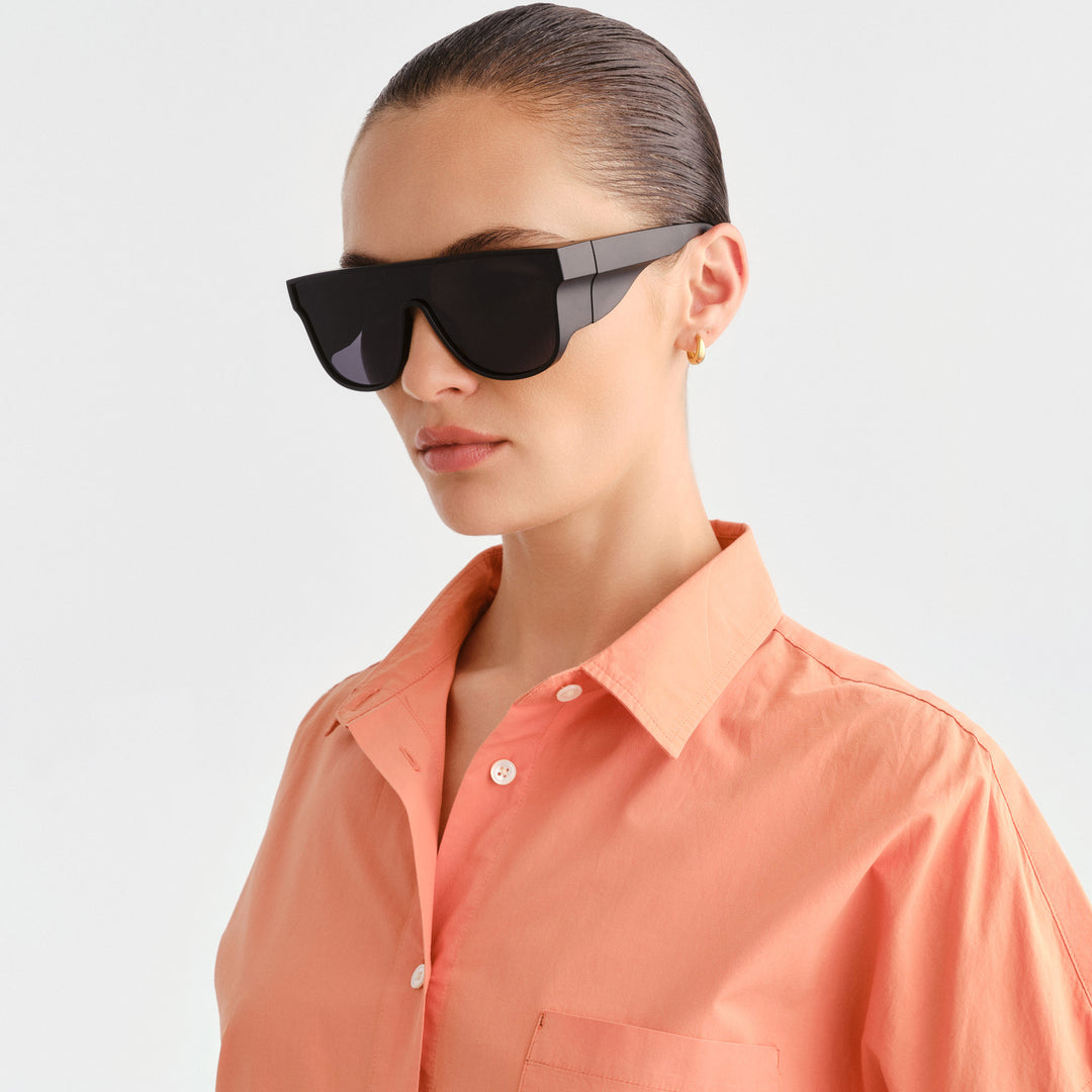 A model wearing oversized black sunglasses with a glossy black temple and a coral pink button-down shirt, photographed against a clean light gray background.