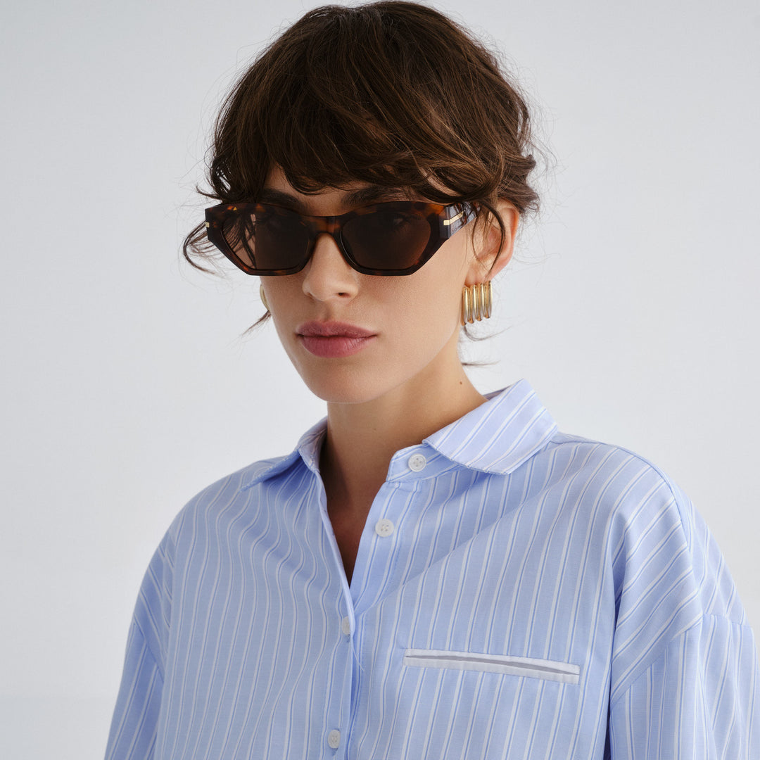 Woman wearing a light blue striped shirt with sunglasses and hoop earrings on a plain background